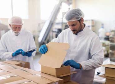 Young Caucasian employee in sterile uniform packing goods in boxes. In background supervisor holding tablet and counting boxes.