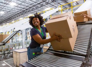 A grinning woman collecting boxes at the bottom of a sorting chute and pushing them along an extendable conveyor belt towards a truck loading dock for delivery.